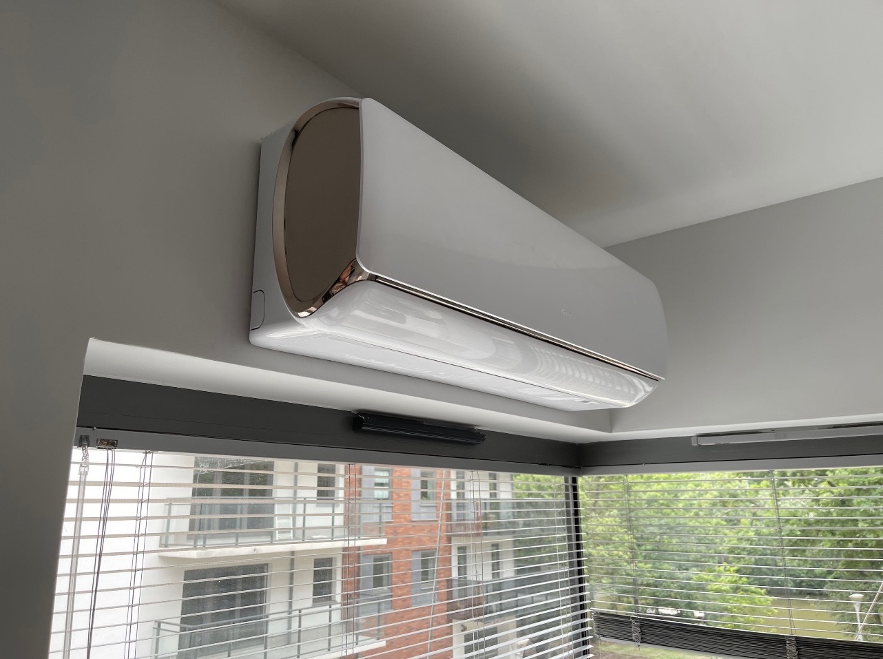 Nowoczesny, biały klimatyzator z chromowanymi detalami zamontowany na szarej ścianie nad oknem z żaluzjami, widok na budynek mieszkalny i zieleń za oknem.