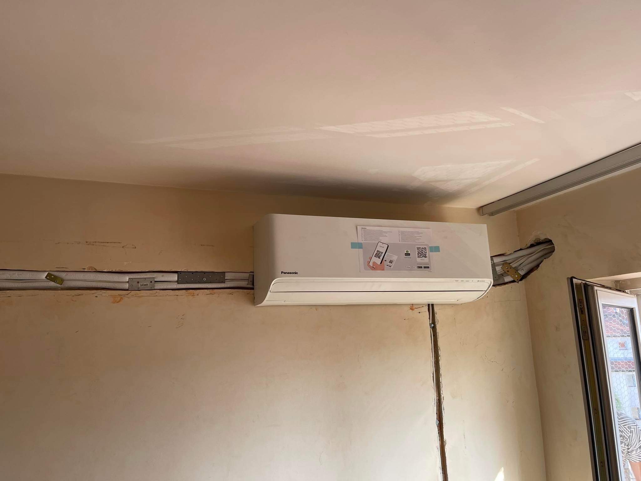 Zamontowana jednostka wewnętrzna klimatyzacji Panasonic na ścianie, z widocznymi przewodami i instalacją w trakcie prac wykończeniowych. Na urządzeniu naklejka z kodami QR.