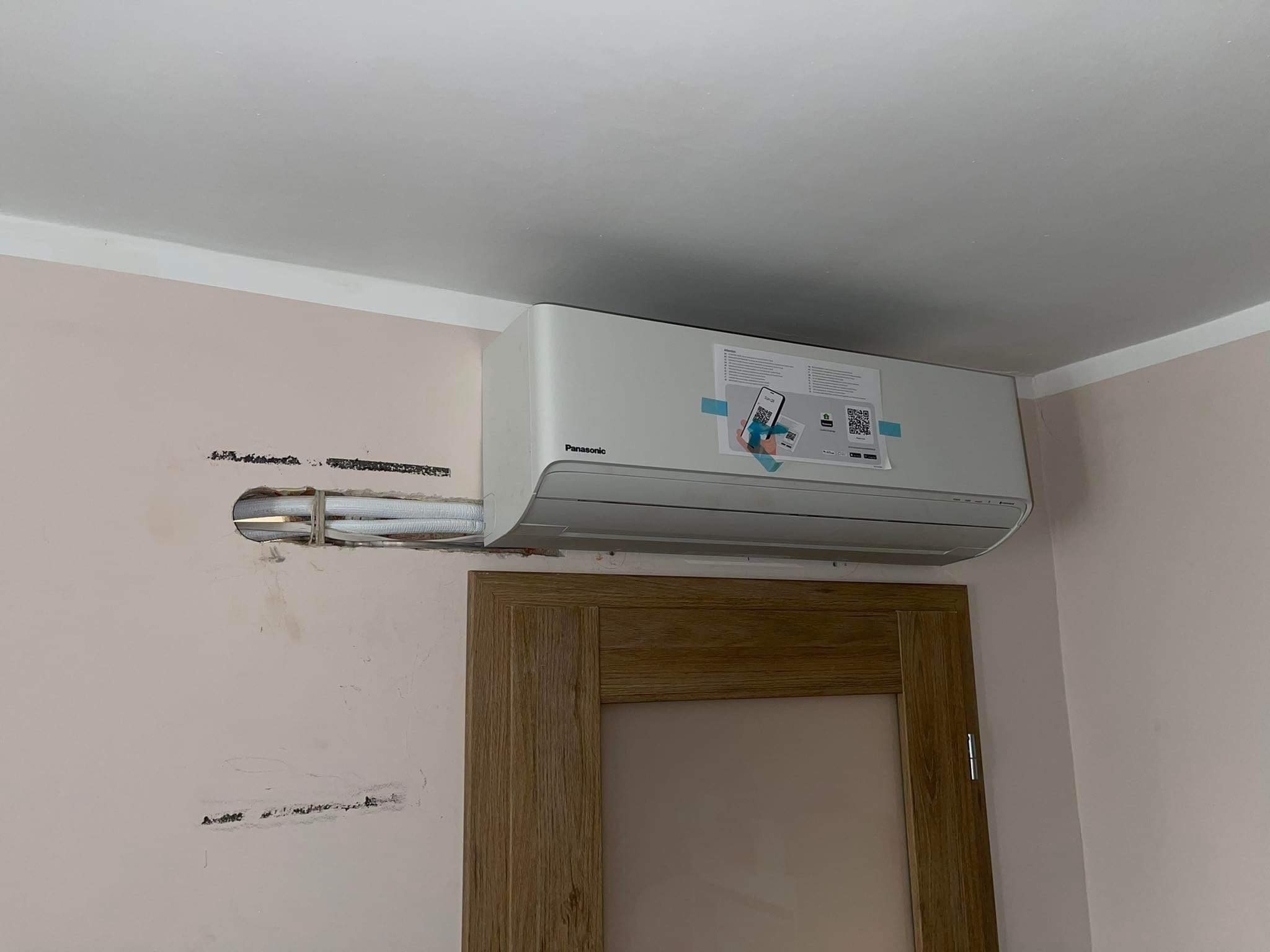 Zamontowana jednostka wewnętrzna klimatyzacji Panasonic na ścianie nad drewnianą ościeżnicą drzwiową, widoczne przewody wychodzące ze ściany przed zamaskowaniem.