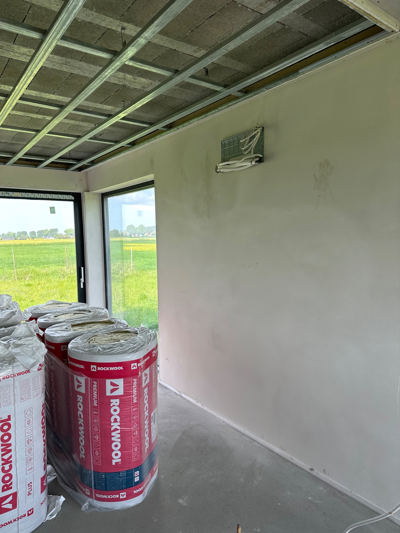 Wnętrze w trakcie prac wykończeniowych: widoczne rolki wełny mineralnej Rockwool, metalowa konstrukcja sufitu podwieszanego oraz przygotowane miejsce na instalację klimatyzacji na ścianie...
