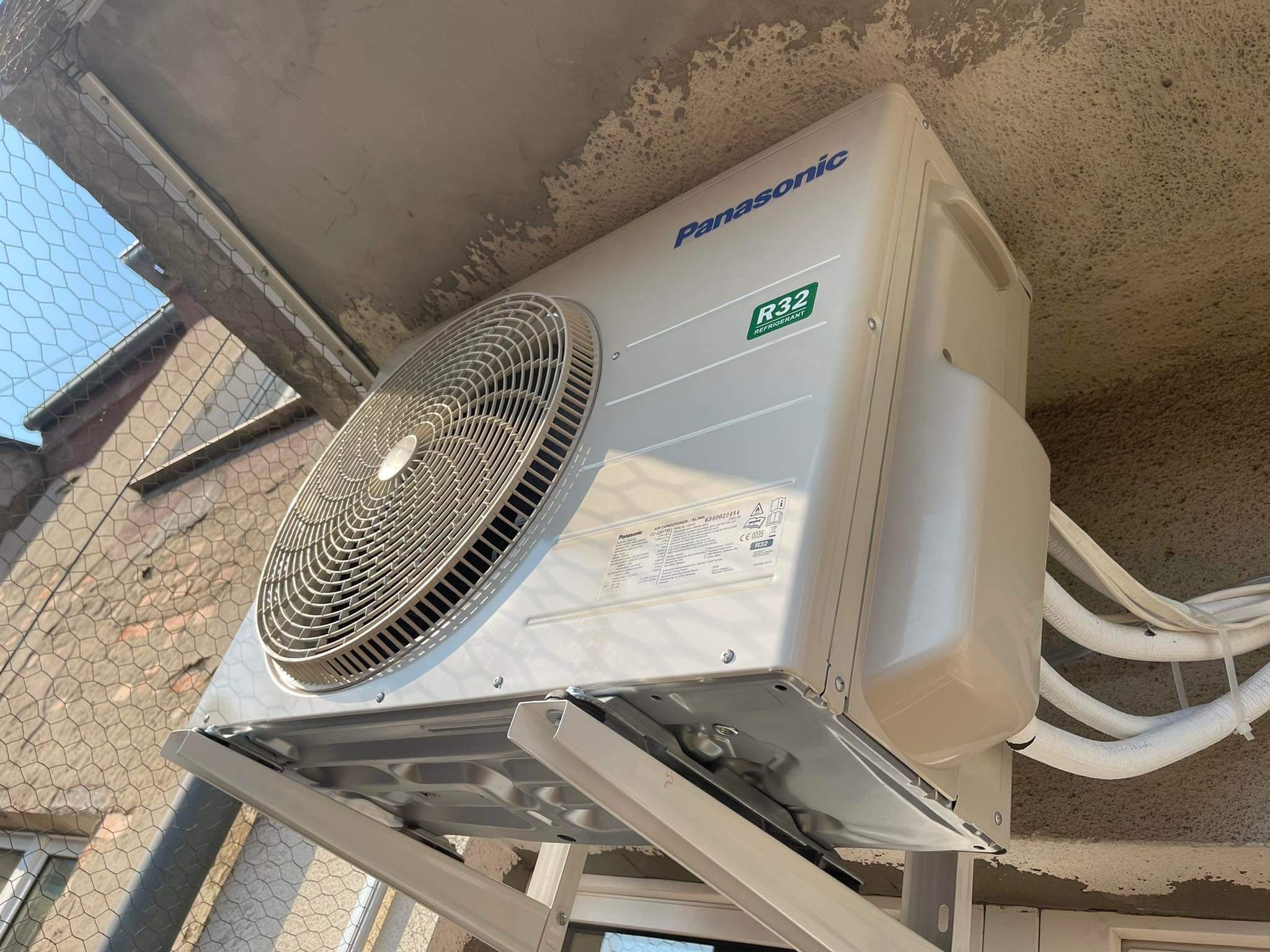 Zamontowana jednostka zewnętrzna klimatyzacji Panasonic R32 na balkonie we Wrocławiu, widok od dołu na wsporniki i rury chłodnicze, siatka zabezpieczająca balkon w tle.