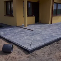 Nowo ułożony taras z szarej kostki brukowej z ciemnym obrzeżem przy żółtym domu. Na piasku leżą narzędzia i fragmenty kostki.