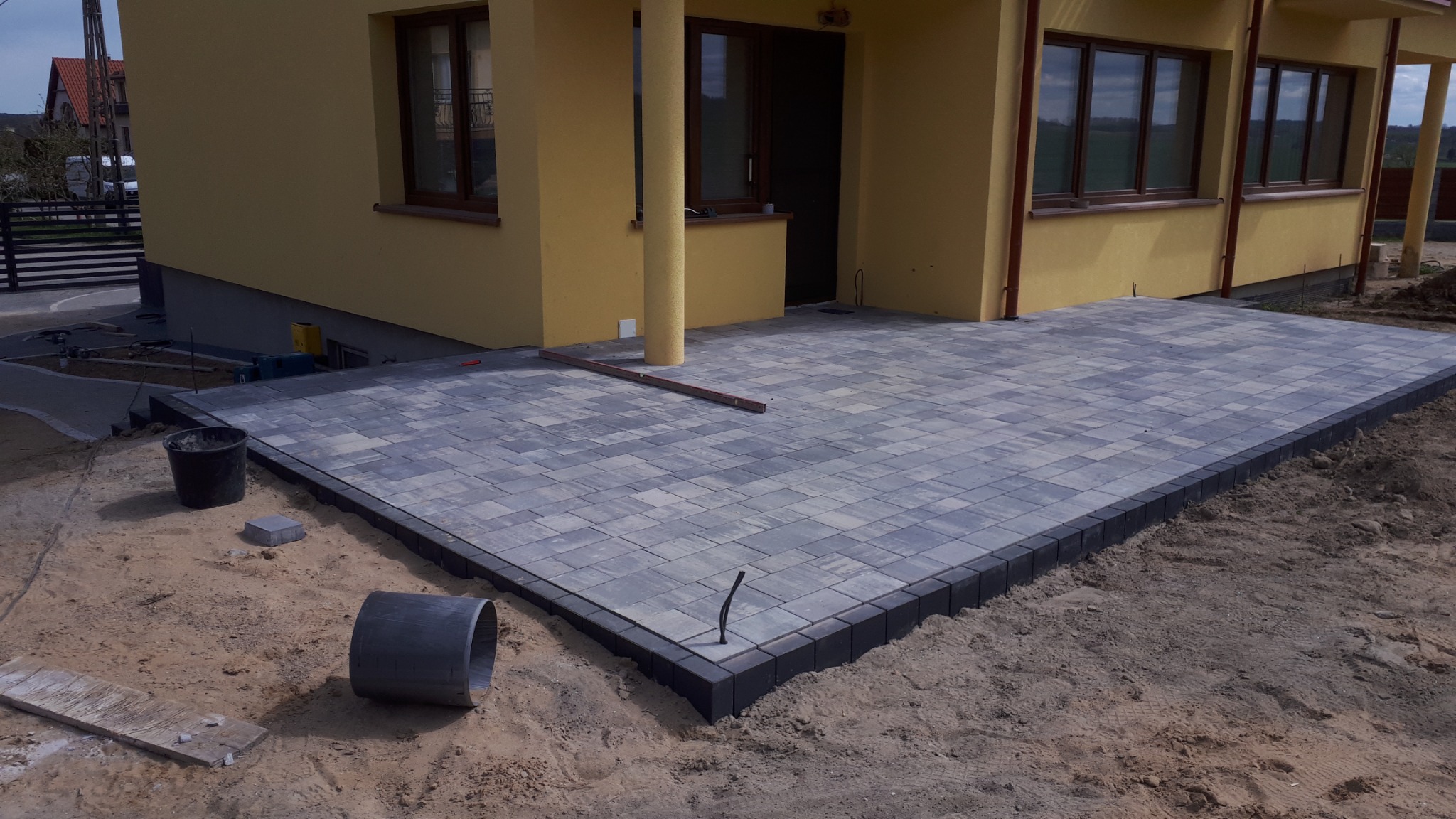 Nowo ułożony taras z szarej kostki brukowej z ciemnym obrzeżem przy żółtym domu. Na piasku leżą narzędzia i fragmenty kostki.