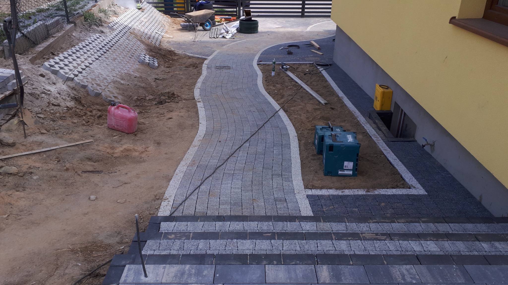 Ścieżka z kostki brukowej w trakcie budowy, z widocznymi narzędziami i materiałami, prowadząca od schodów do domu z żółtą elewacją, obok przygotowane podłoże pod dalsze prace.