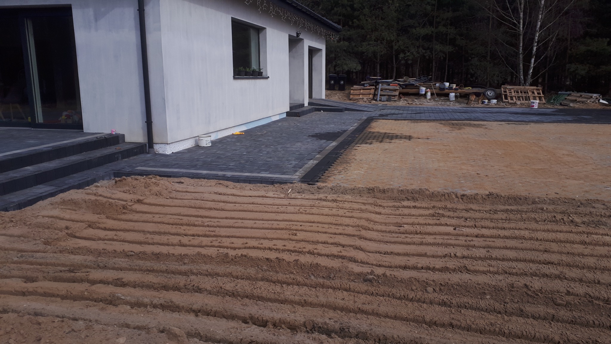 Układanie kostki brukowej przy domu jednorodzinnym, widoczne przygotowane podłoże z wyprofilowanymi śladami, częściowo ułożona kostka, materiały budowlane w tle.