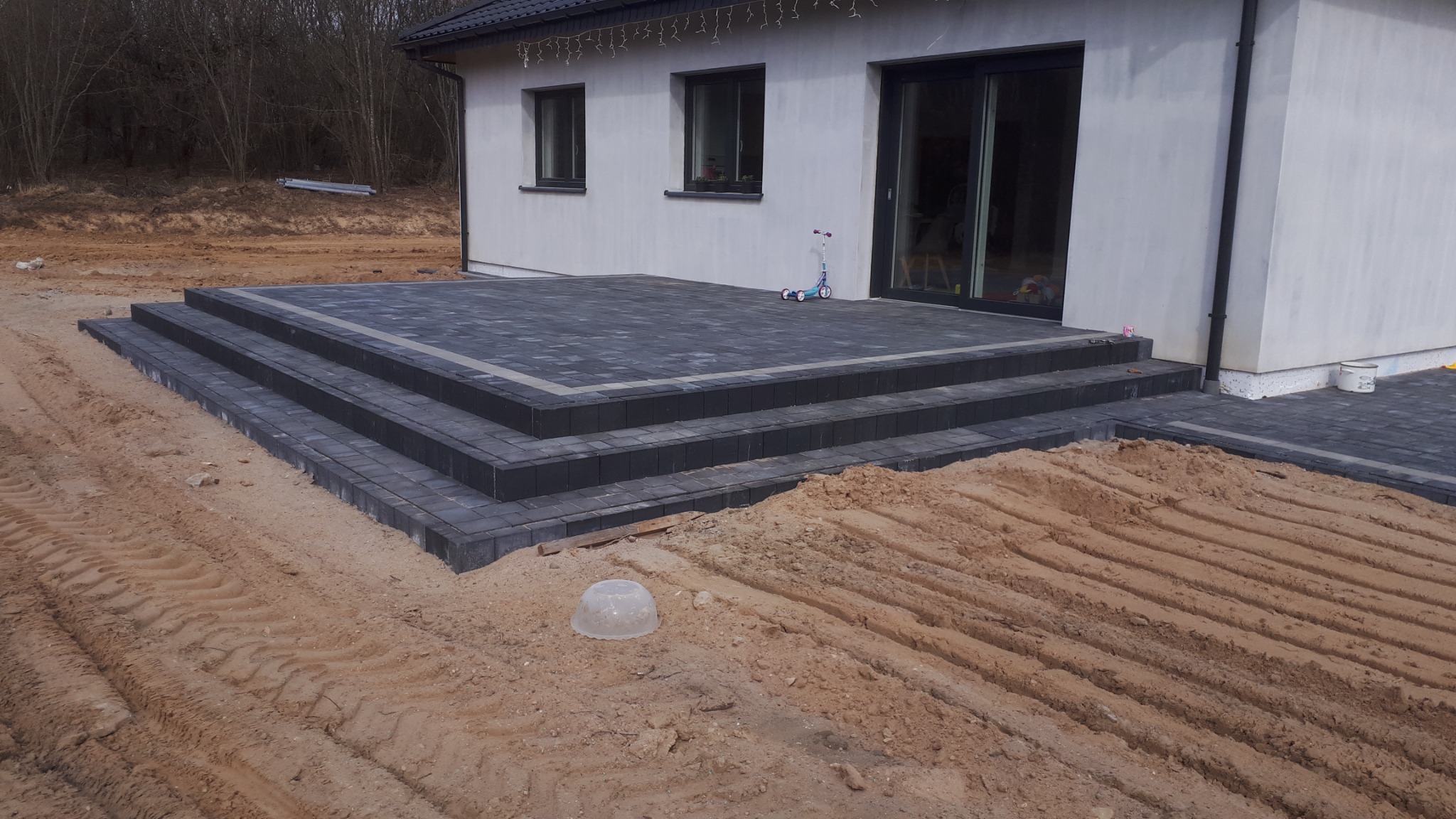 Nowoczesny taras z kostki brukowej w odcieniach szarości, z trzema stopniami wejściowymi, częściowo otoczony piaskiem i ziemią, z hulajnogą dziecięcą zaparkowaną obok drzwi tarasowych.