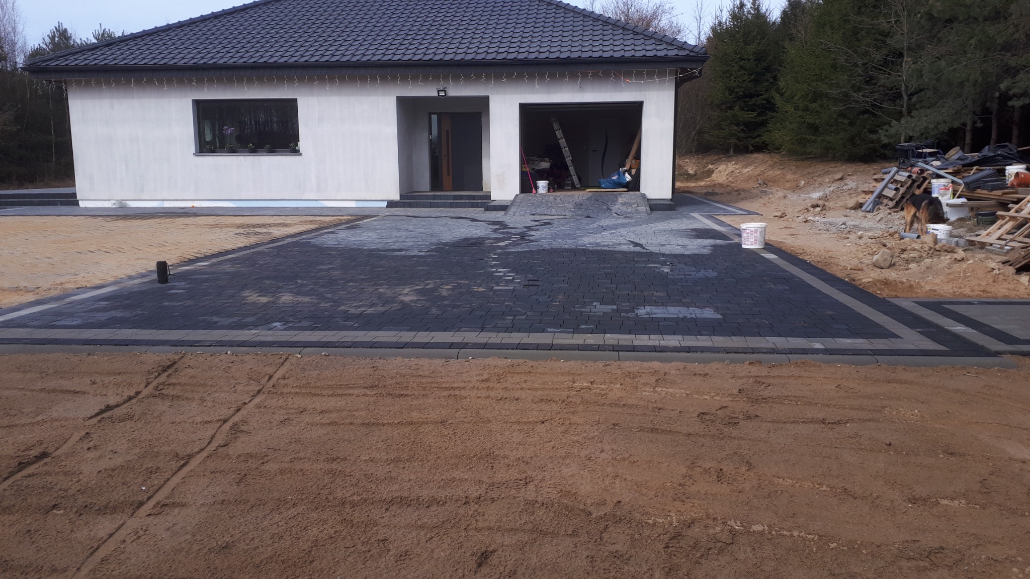 Nowo ułożona, ciemnoszara kostka brukowa z jasnoszarą obwódką przed nowoczesnym domem z ciemnym dachem; widoczne ślady po oponach na piasku, pies rasy owczarek niemiecki w tle.