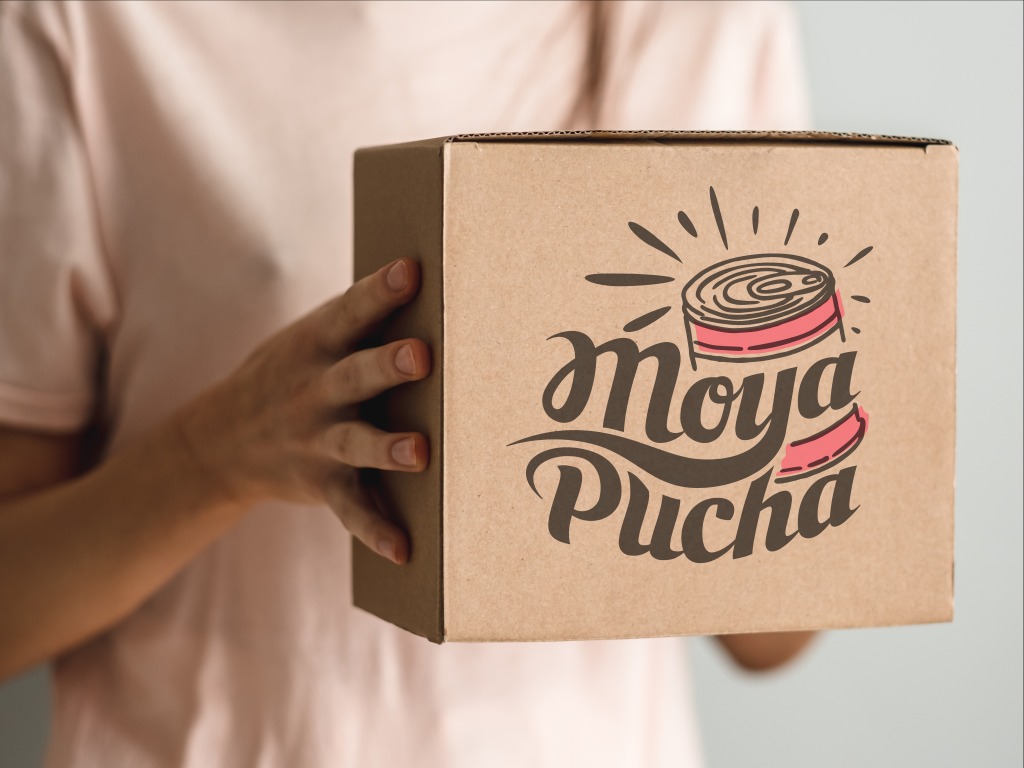 Osoba trzyma brązowe pudełko z nadrukowanym logo 'Moya Pucha', przedstawiającym stylizowaną puszkę konserw z różowym wypełnieniem.