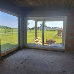 IWMAR IWONA ŁASKA - Surowy stan: Zamontowane okno przesuwne w murze z widokiem na zewnątrz. Palety w tle, betonowa podłoga i surowy strop. Wnętrze w trakcie budowy.