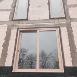 IWMAR IWONA ŁASKA - Montaż okien na surowej ścianie budynku. Widoczne okno przesuwne i dwa wąskie okna pionowe na wyższych kondygnacjach. W oknach odbijają się drzewa.