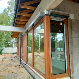 Surowy stan budowy: montaż dużych, drewnopodobnych okien z roletami zewnętrznymi w nowym budynku z surowymi ścianami, widoczne elementy konstrukcyjne dachu i izolacja fundamentów.