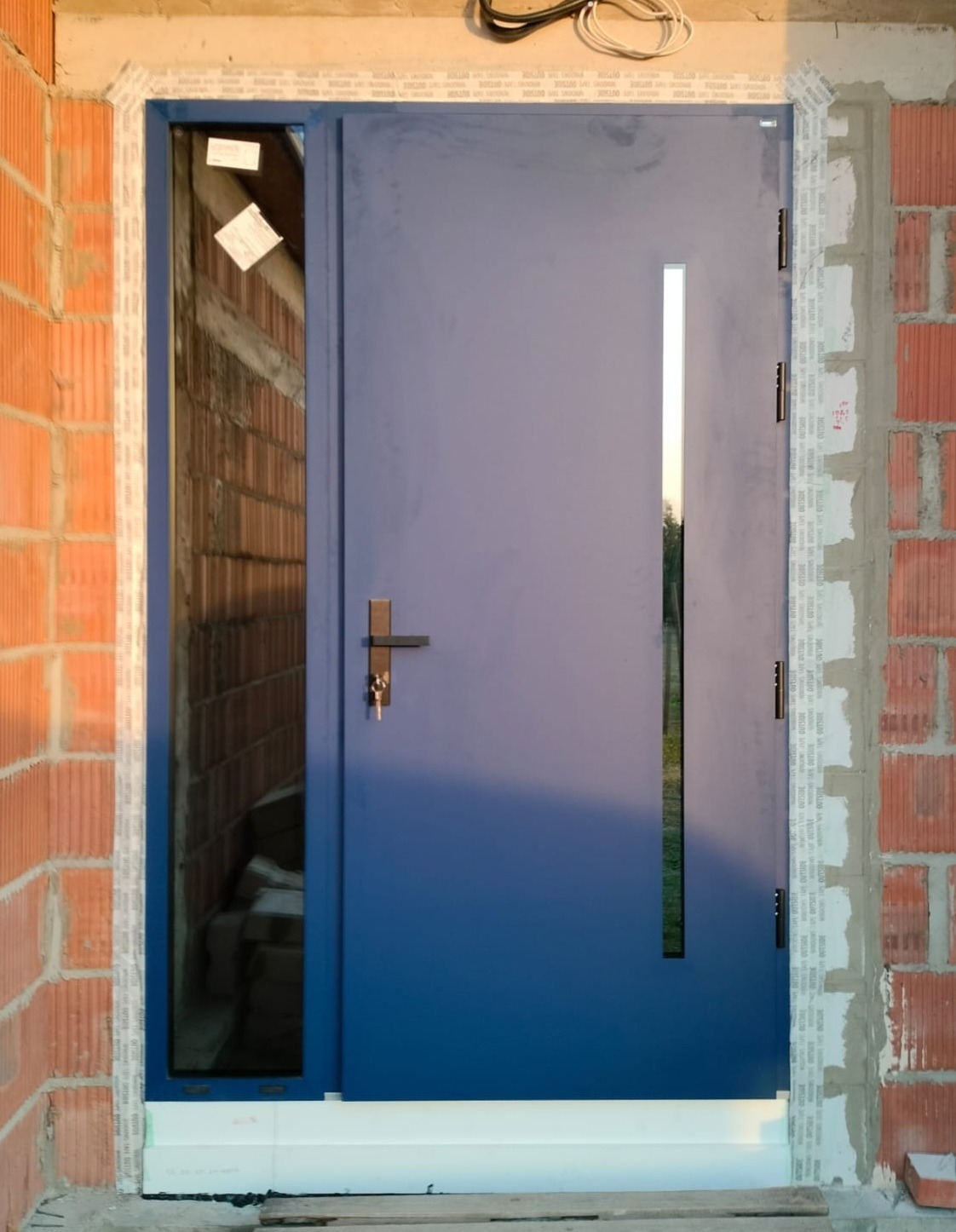 Niebieskie drzwi wejściowe z wąskim przeszkleniem bocznym, zamontowane w surowym murze. Widoczne taśmy uszczelniające i fragmenty cegieł. Nowoczesny design.