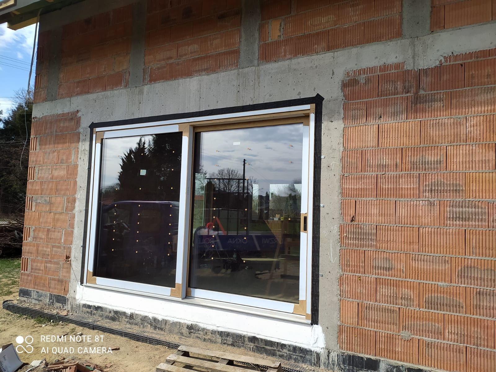 Montaż okna przesuwnego w surowym murze z cegły kratówki. Widoczne uszczelnienia i elementy montażowe. W szybie odbija się otoczenie.