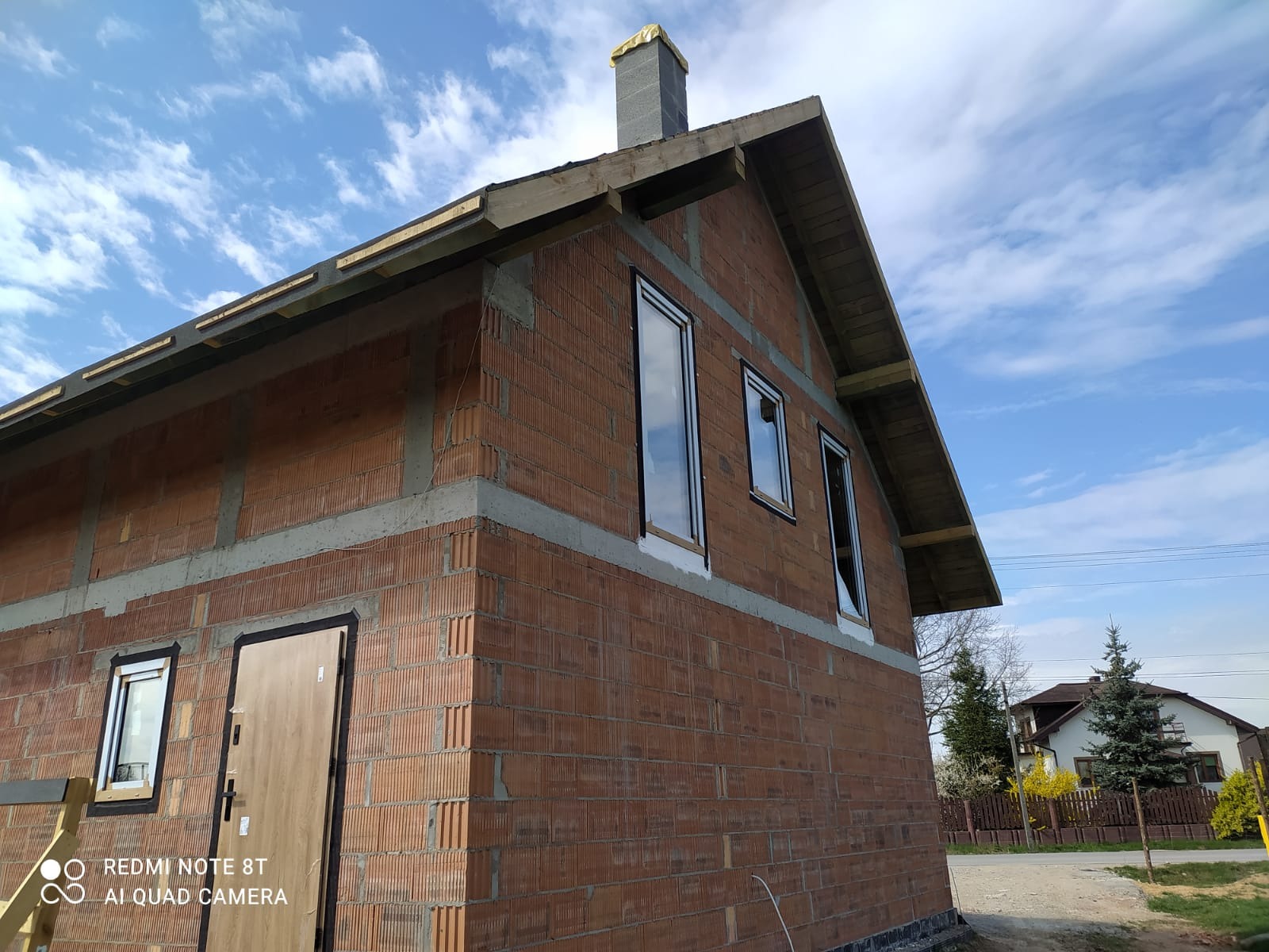 Surowy budynek z cegły z zamontowanymi oknami i drzwiami, widok z zewnątrz w słoneczny dzień. Budynek w trakcie budowy, widoczne elementy konstrukcyjne dachu.