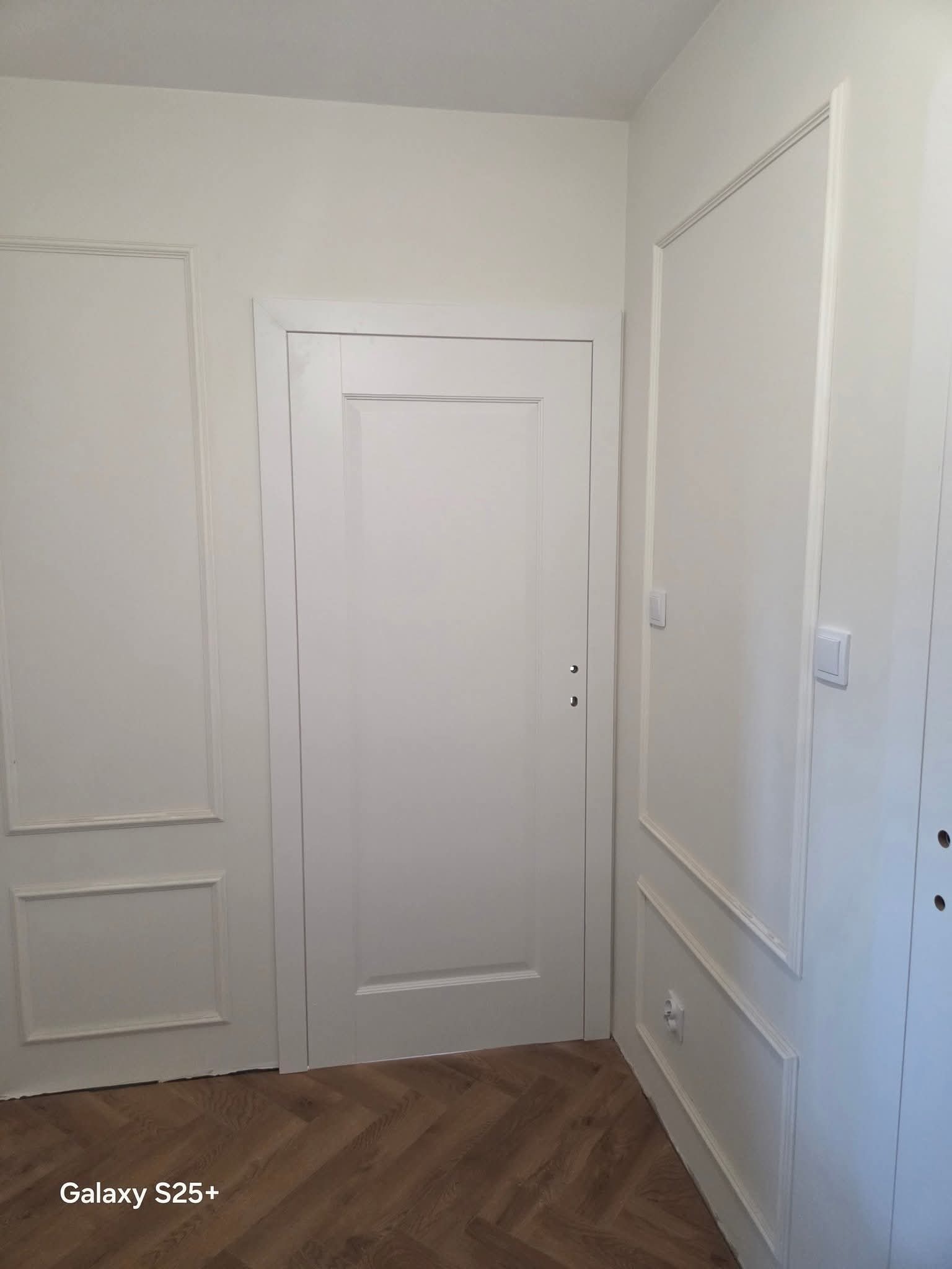 Białe drzwi wewnętrzne z ozdobnymi listwami na ścianach, parkiet w jodełkę. Minimalistyczny design z subtelnymi detalami.