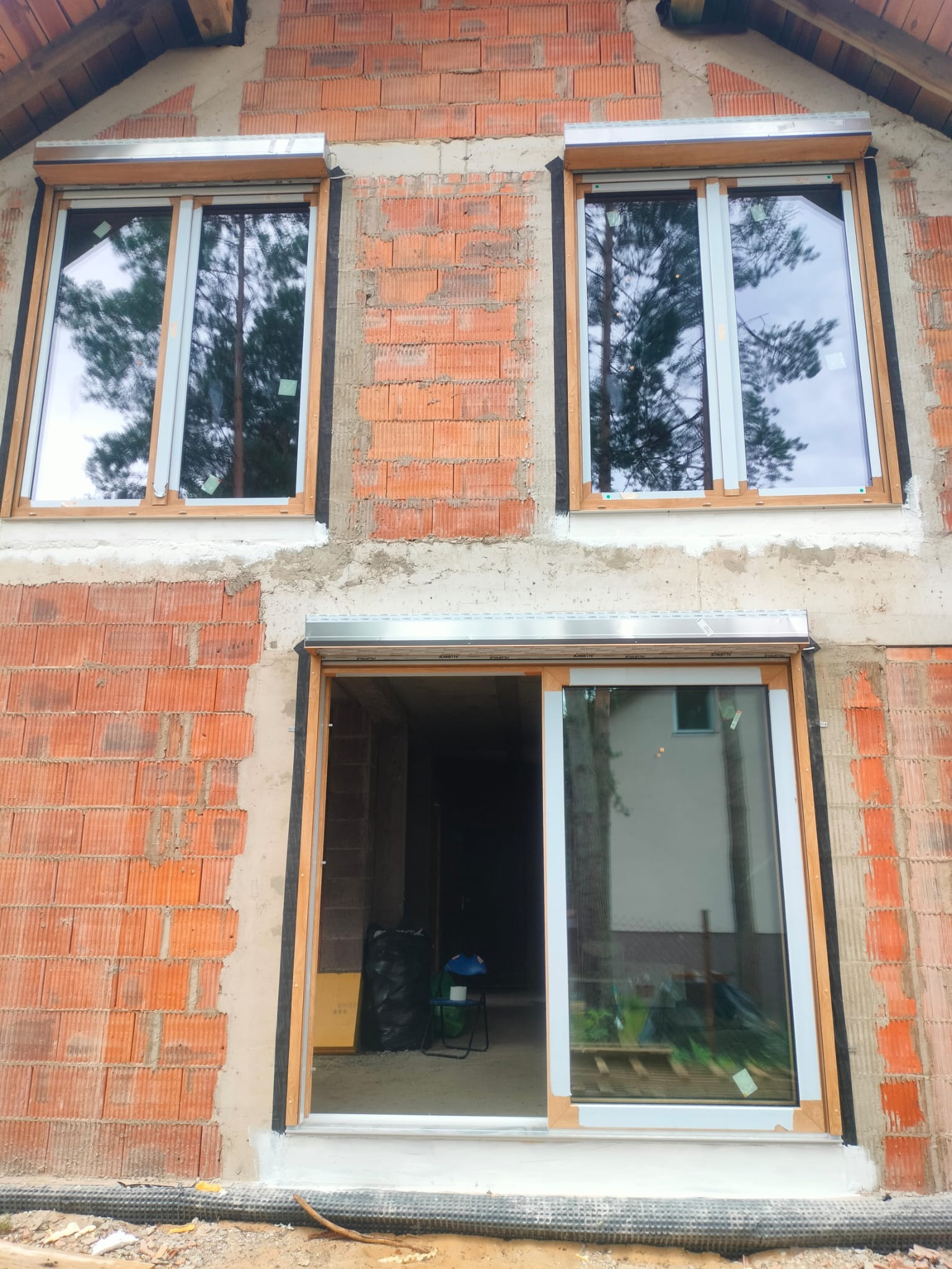 Surowy mur z oknami z roletami w trakcie montażu. Widoczne niedokończone prace murarskie i materiały budowlane. W oknach odbija się otoczenie.