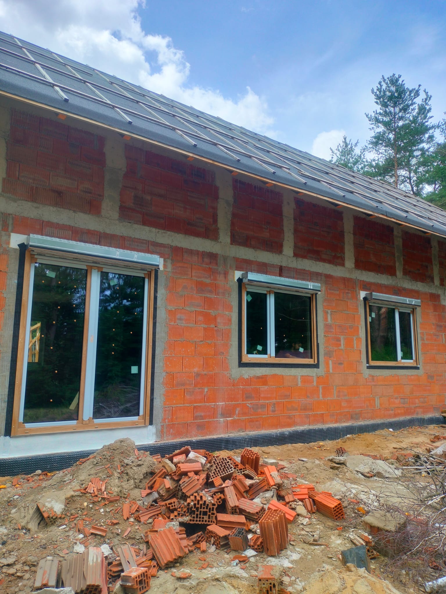 Budynek w trakcie budowy z zamontowanymi oknami z roletami zewnętrznymi, widoczne niedokończone ściany z cegły i gruz. Ujęcie z dołu, w tle fragment lasu.