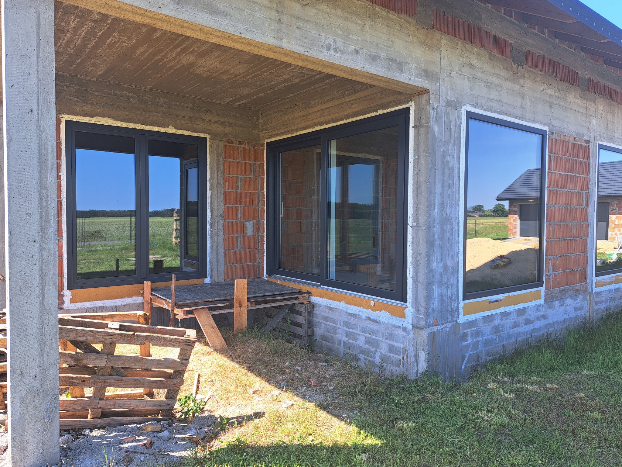 Montaż okien PCV z czarną ramą w surowym budynku z cegły. Widoczne rusztowania i przygotowania do dalszych prac wykończeniowych. Okna odbijają krajobraz.