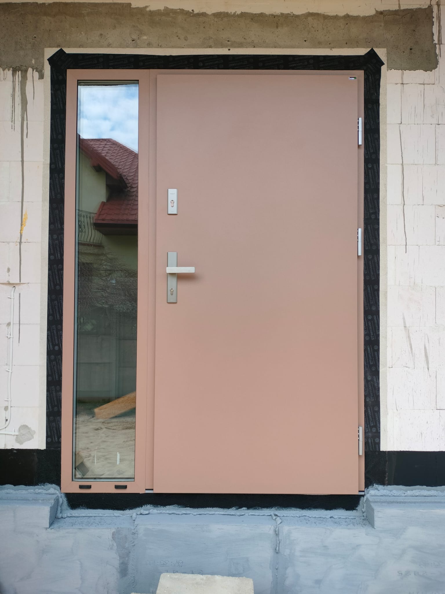 Zewnętrzne drzwi wejściowe z szybą boczną, zamontowane w surowym stanie budynku. Widoczne uszczelnienia i taśmy izolacyjne. W szybie odbija się fragment budynku.