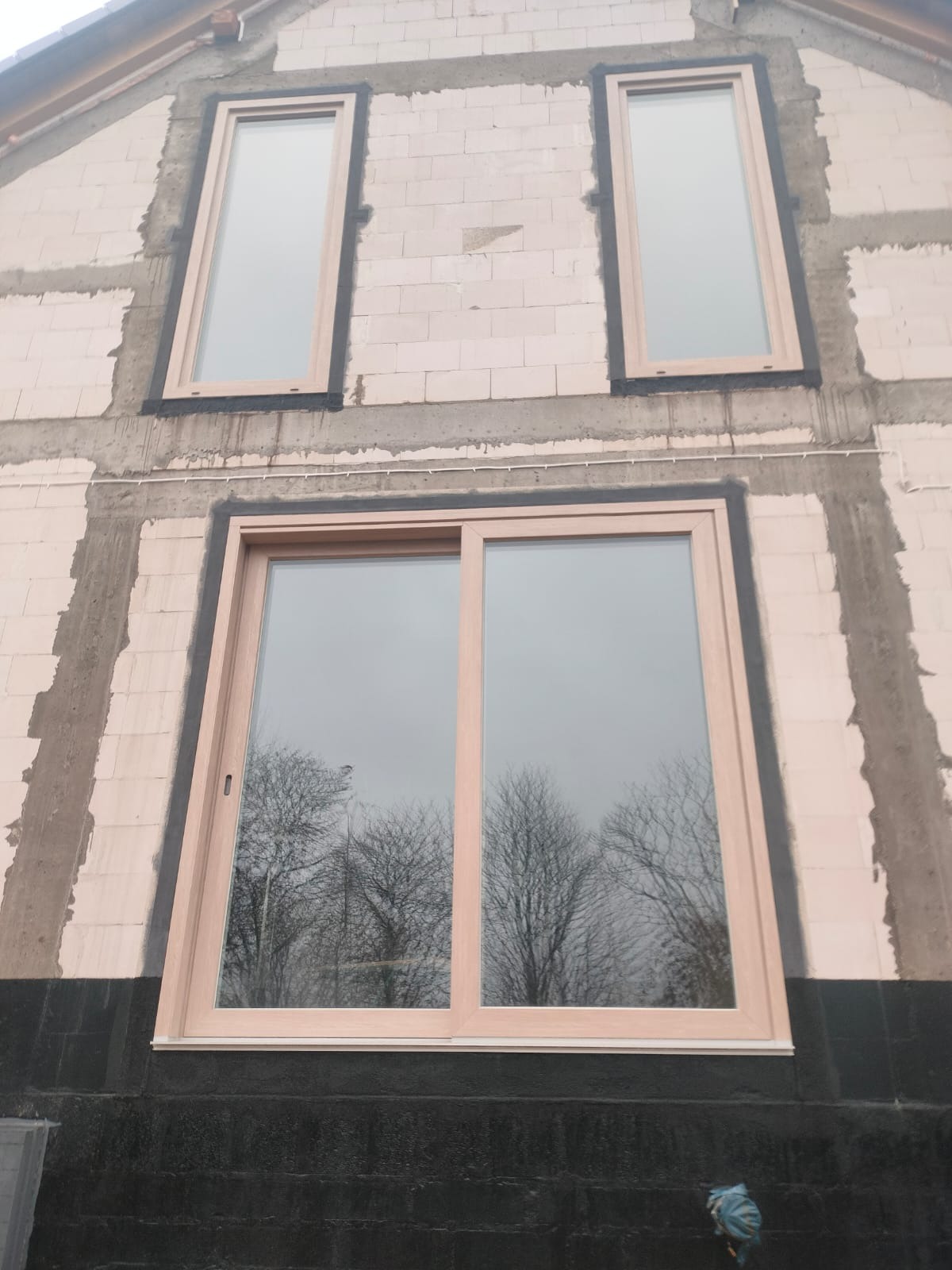 Montaż okien na surowej ścianie budynku. Widoczne okno przesuwne i dwa wąskie okna pionowe na wyższych kondygnacjach. W oknach odbijają się drzewa.