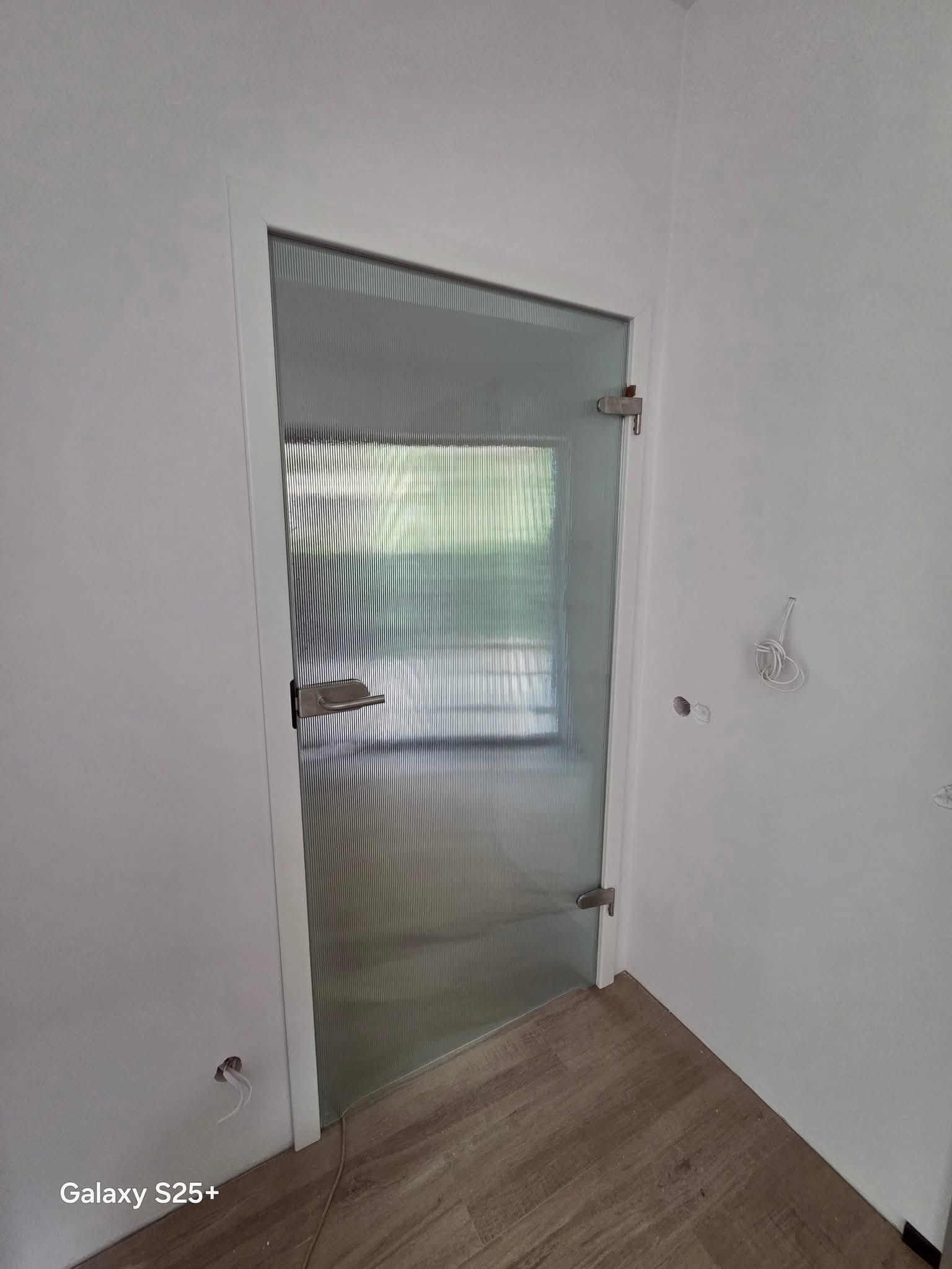 Szklane drzwi wewnętrzne z ryflowanym szkłem i chromowanym uchwytem, zamontowane w białej futrynie, widoczne na tle jasnych ścian i drewnopodobnej podłogi.