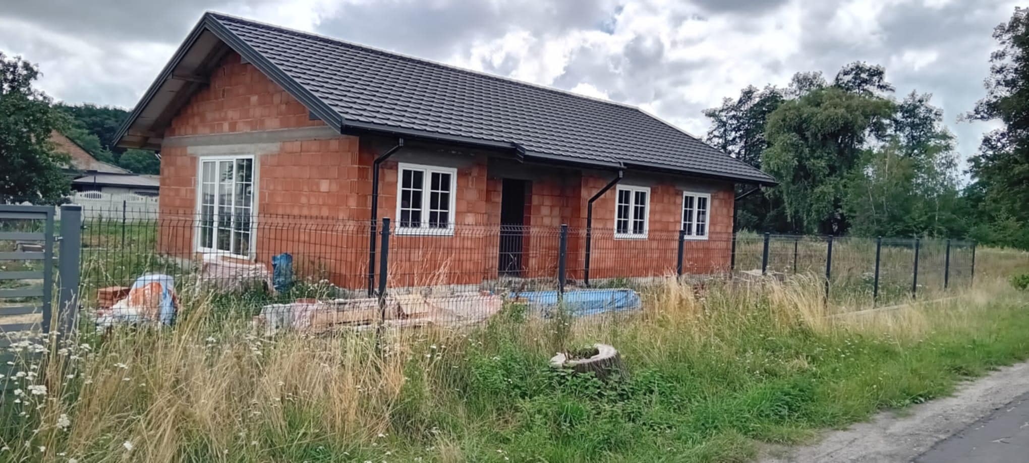 Nowy dom w trakcie budowy z zamontowanymi oknami PCV, ogrodzony siatką. Widoczne materiały budowlane i wysoka trawa. Dach pokryty dachówką. Pochmurne niebo w tle.