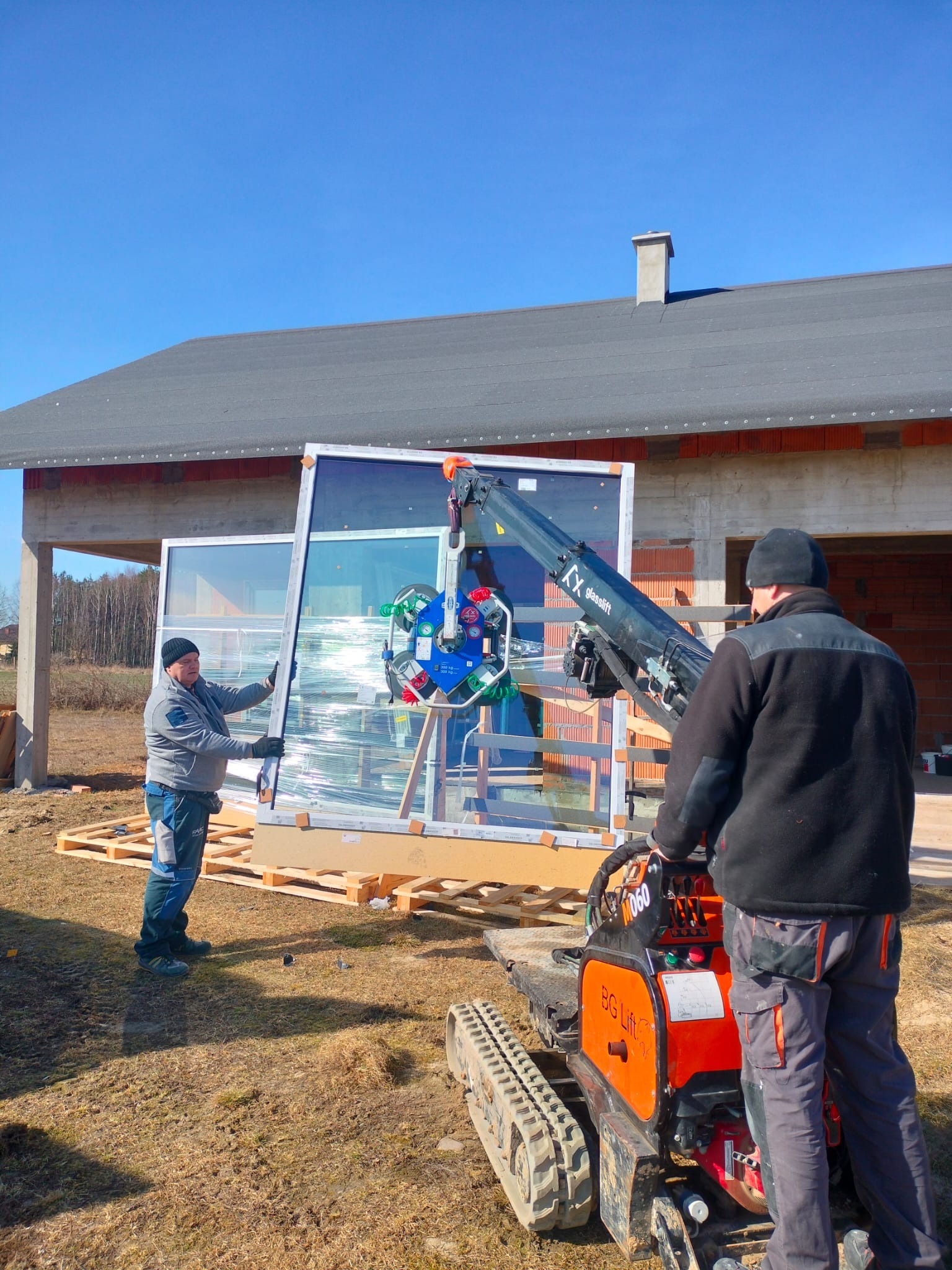 Montaż okna z użyciem mini żurawia przyssawkowego Glaslift na placu budowy. Dwie osoby nadzorują proces. W tle widoczny budynek w stanie surowym otwartym.