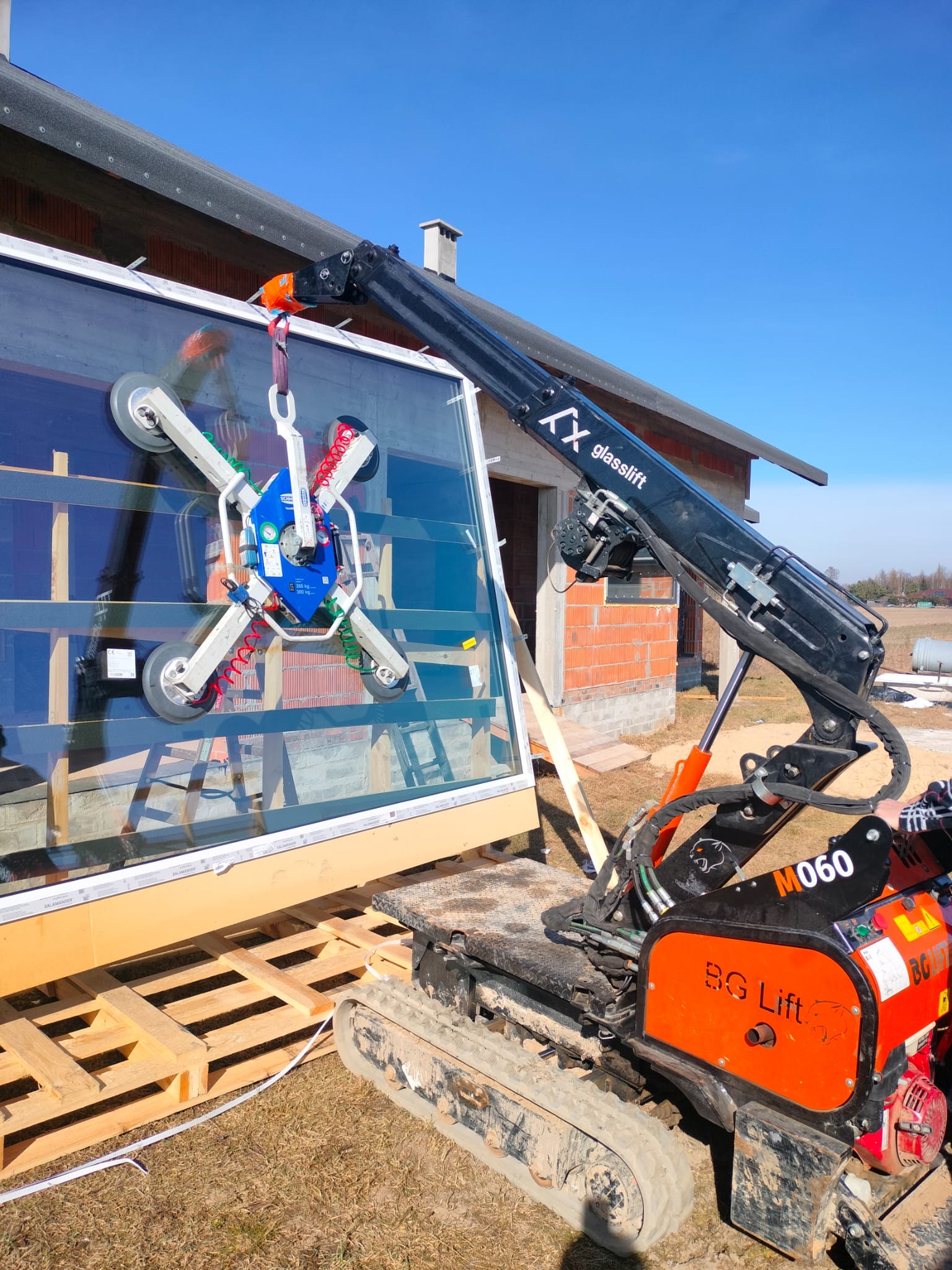 Montaż okna z użyciem przyssawki i mini-żurawia BG Lift na tle surowego budynku. Widoczne detale mocowania i profil okienny Salamander.