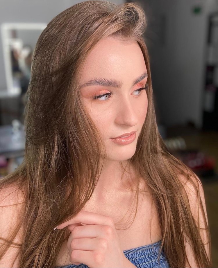 Portret kobiety z delikatnym, brzoskwiniowym cieniem na powiekach i błyszczącymi ustami, patrzącej w bok, z dłonią blisko dekoltu.