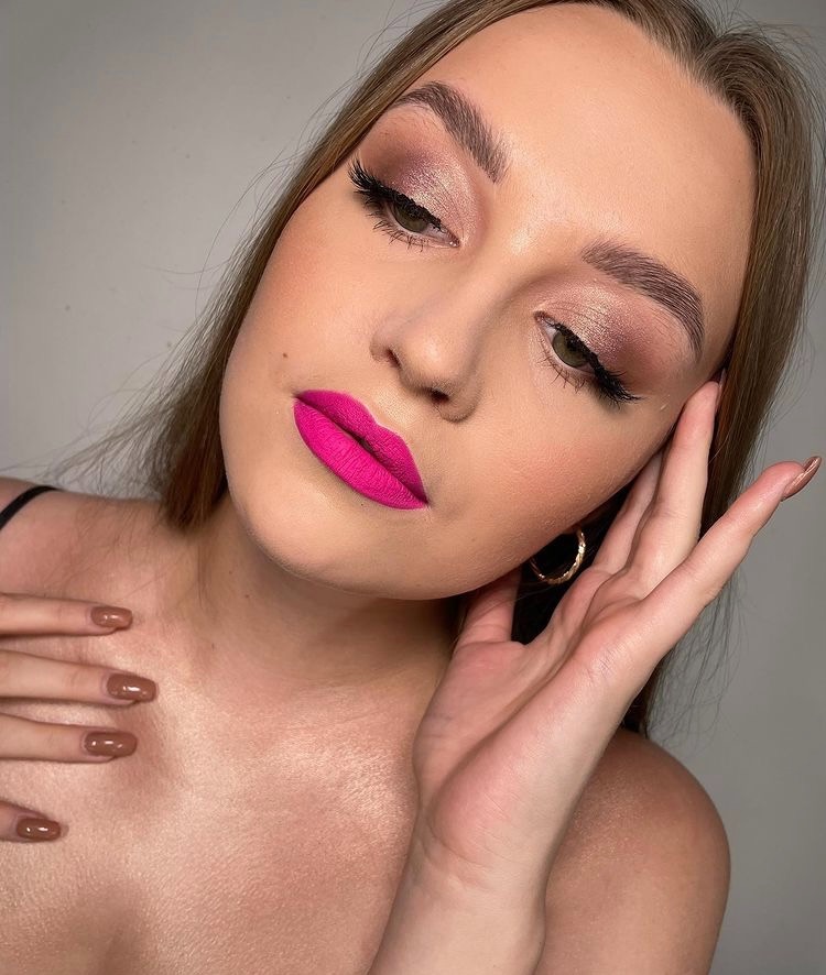 Portret kobiety z intensywnym różem na ustach, subtelnym cieniem do powiek i przedłużonymi rzęsami, dłoń przy twarzy, widoczne złote kolczyki koła i brązowy manicure.