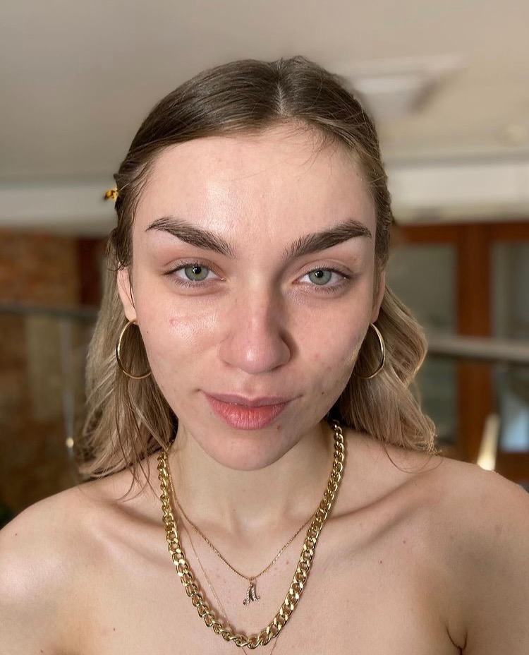 Portret kobiety z blond włosami i zielonymi oczami przed aplikacją kosmetyków, widoczne naturalne brwi przed stylizacją i złota biżuteria.