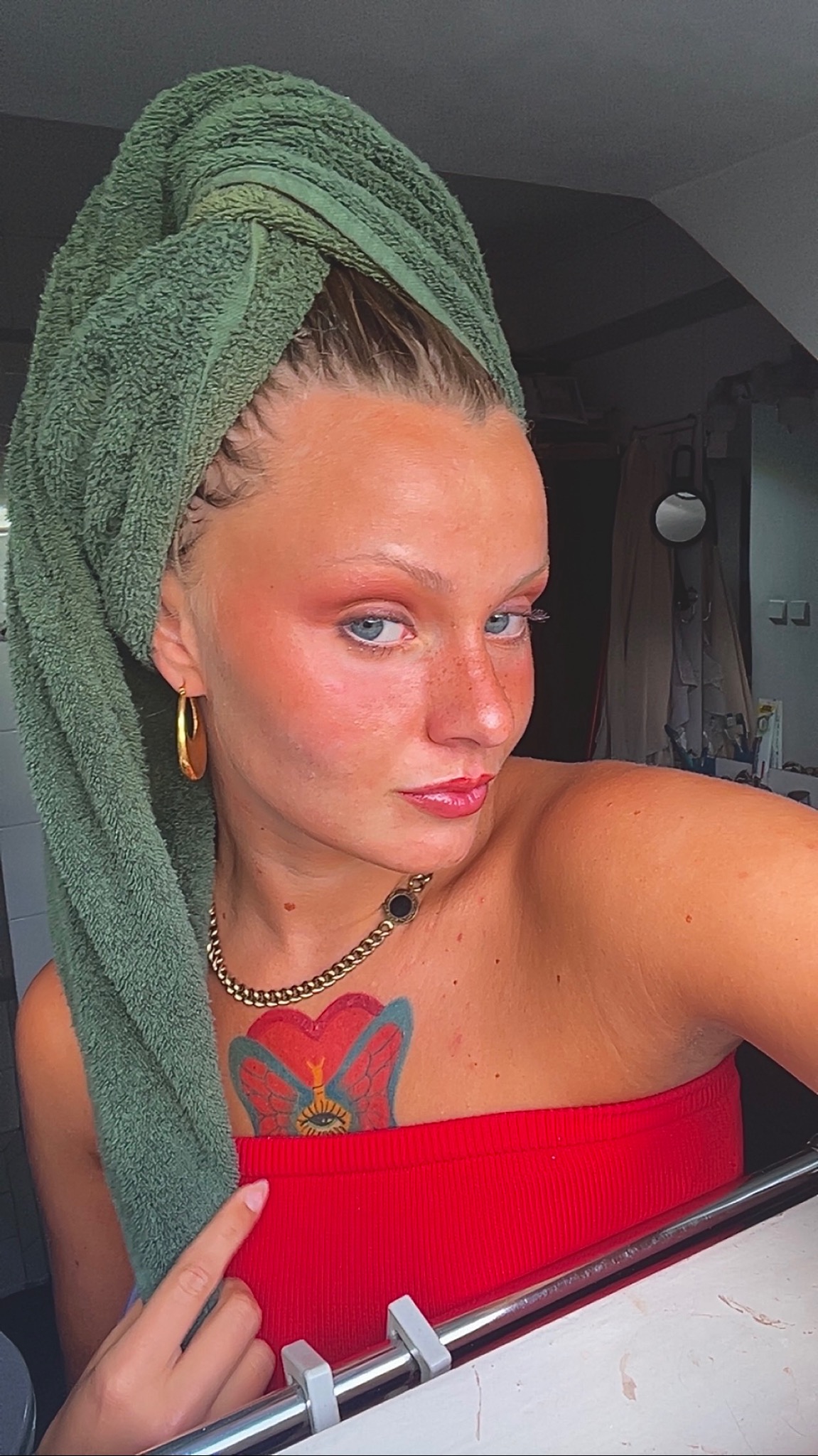 Kobieta z zielonym ręcznikiem na głowie, w czerwonej bluzce i z wyrazistym, różowym rumieńcem oraz pomalowanymi ustami. Widoczny tatuaż na dekolcie i złota biżuteria.