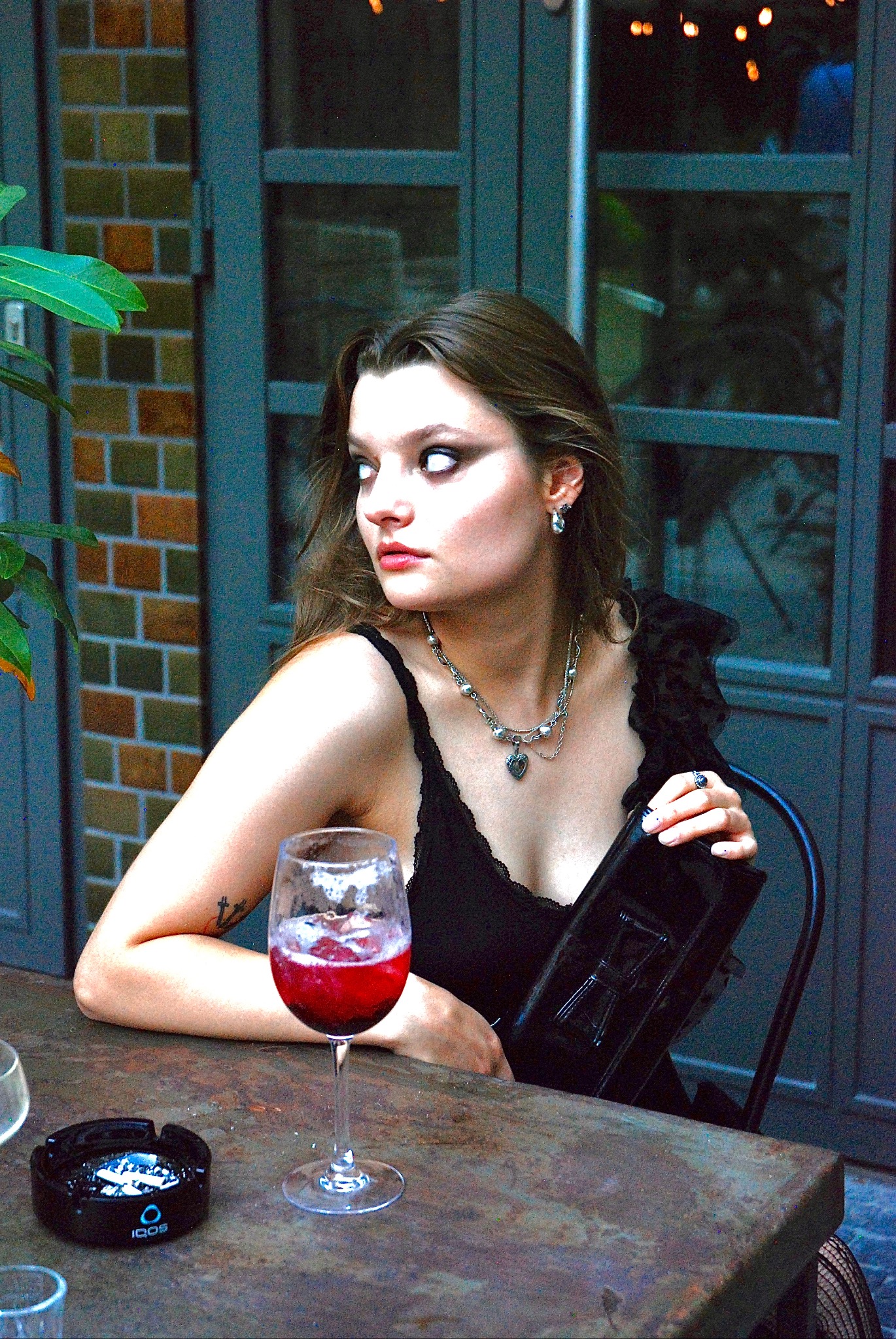 Kobieta z jasną cerą, w czarnej sukience, z ciemnym makijażem oczu i czerwonymi ustami, pozuje przy stoliku z drinkiem. Gotycki styl podkreślają srebrna biżuteria i upięcie włosów.