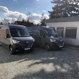 Dwa szare busy Renault Master zaparkowane na żwirowym parkingu przed budynkiem w słoneczny dzień.