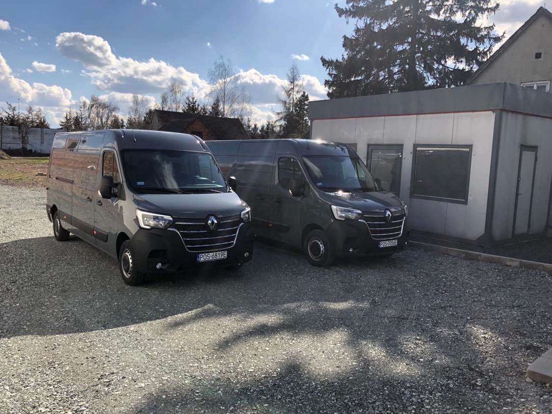 Dwa szare busy Renault Master zaparkowane na żwirowym parkingu przed budynkiem w słoneczny dzień.