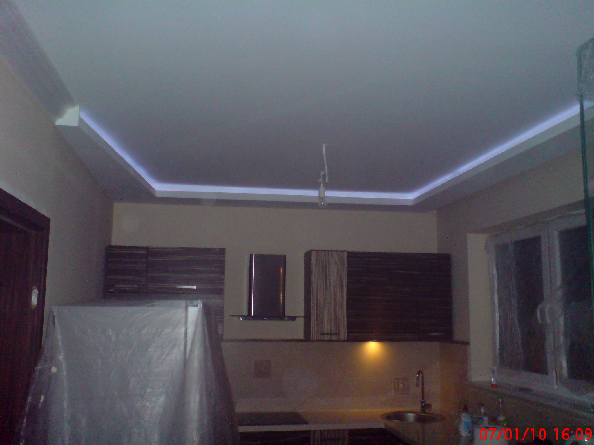 Sufit podwieszany z oświetleniem LED w kuchni, widoczna zabudowa meblowa i zakryta folią lodówka.