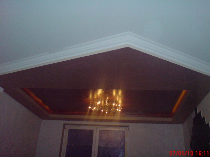 Wykończony sufit podwieszany z płyt gipsowo-kartonowych, pomalowany na brązowo, z ozdobnym oświetleniem i listwami przysufitowymi, widok z dołu na tle okna.