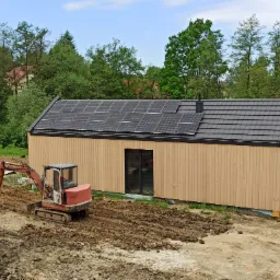 Nowoczesny dom z drewnianą elewacją i panelami słonecznymi na dachu, obok niego stoi mała koparka przygotowująca teren.