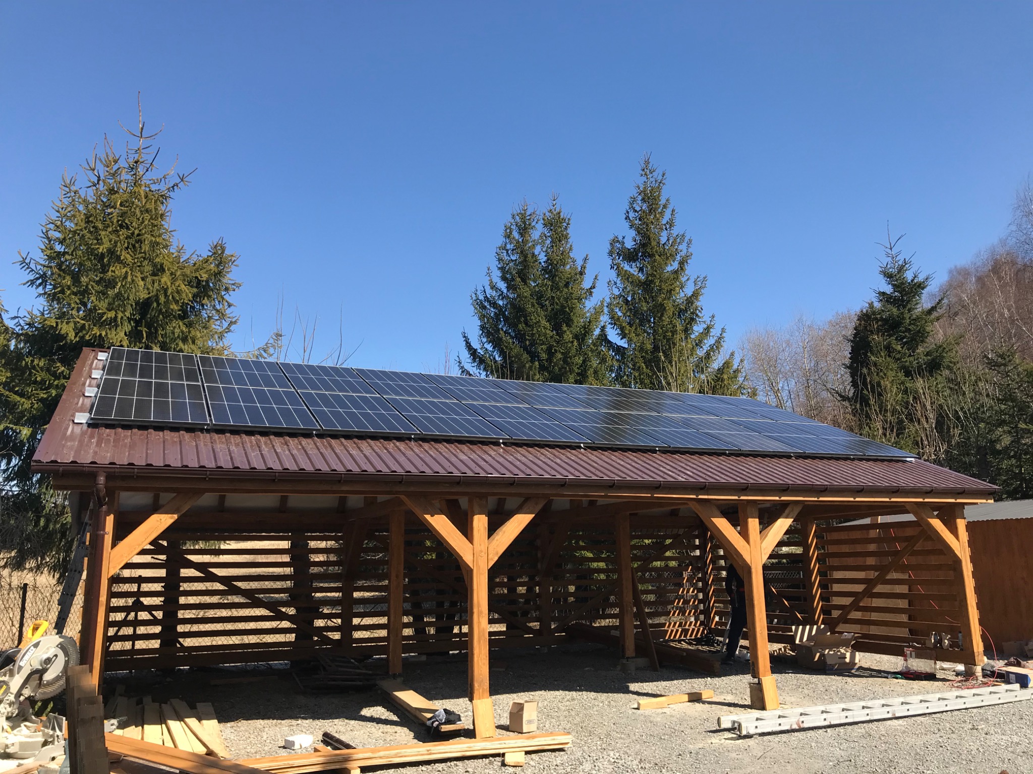 Zadaszona drewniana konstrukcja z brązowym dachem i panelami słonecznymi na tle błękitnego nieba i drzew