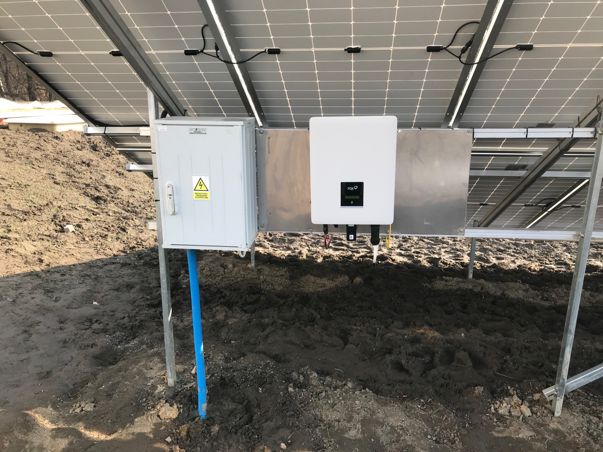 Instalacja paneli słonecznych: widok od spodu na moduły, falownik FOX-D i skrzynkę z zabezpieczeniami, niebieska rura osłonowa kabli w ziemi.