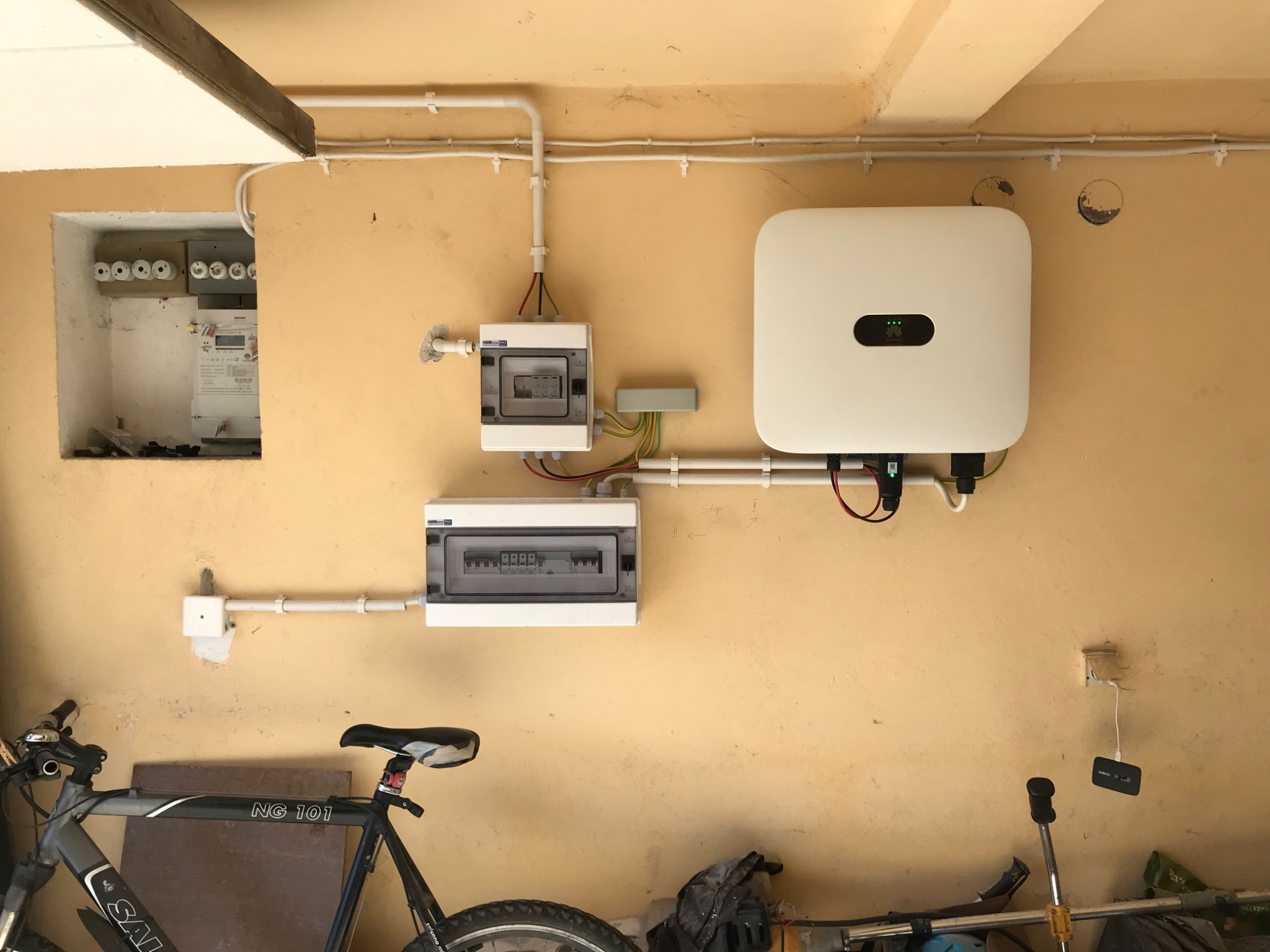 Zamontowany biały falownik fotowoltaiczny Huawei z podłączonymi przewodami, skrzynki elektryczne, bezpieczniki w ścianie oraz rower w garażu.