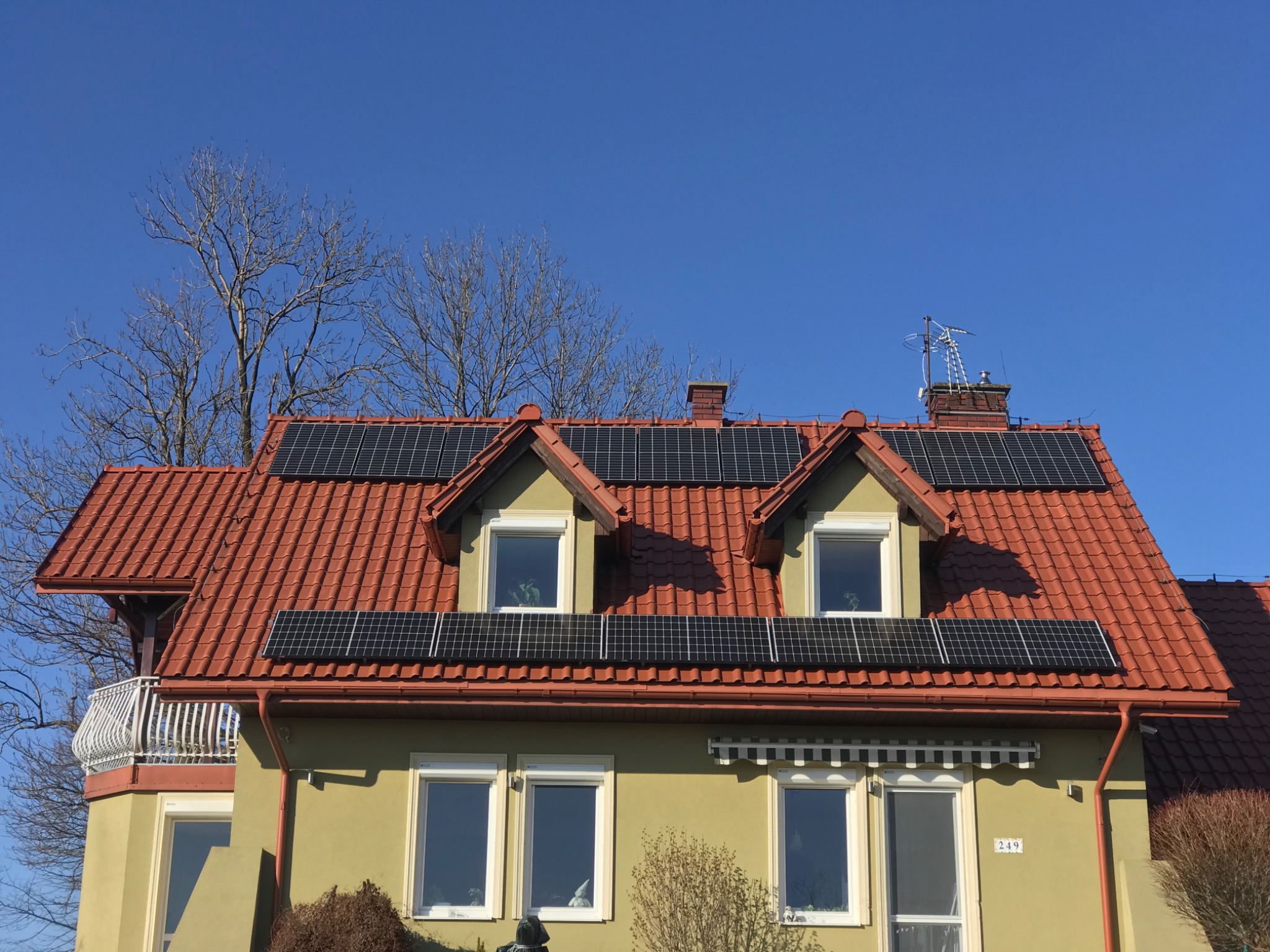 Dom jednorodzinny z panelami słonecznymi na dachu z czerwonej dachówki, widoczne okna dachowe, antena telewizyjna na kominie, bezlistne drzewo na tle błękitnego nieba.