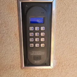 Panel domofonu ACO z klawiaturą numeryczną i wyświetlaczem LCD, zamontowany na beżowej ścianie o fakturze baranka.