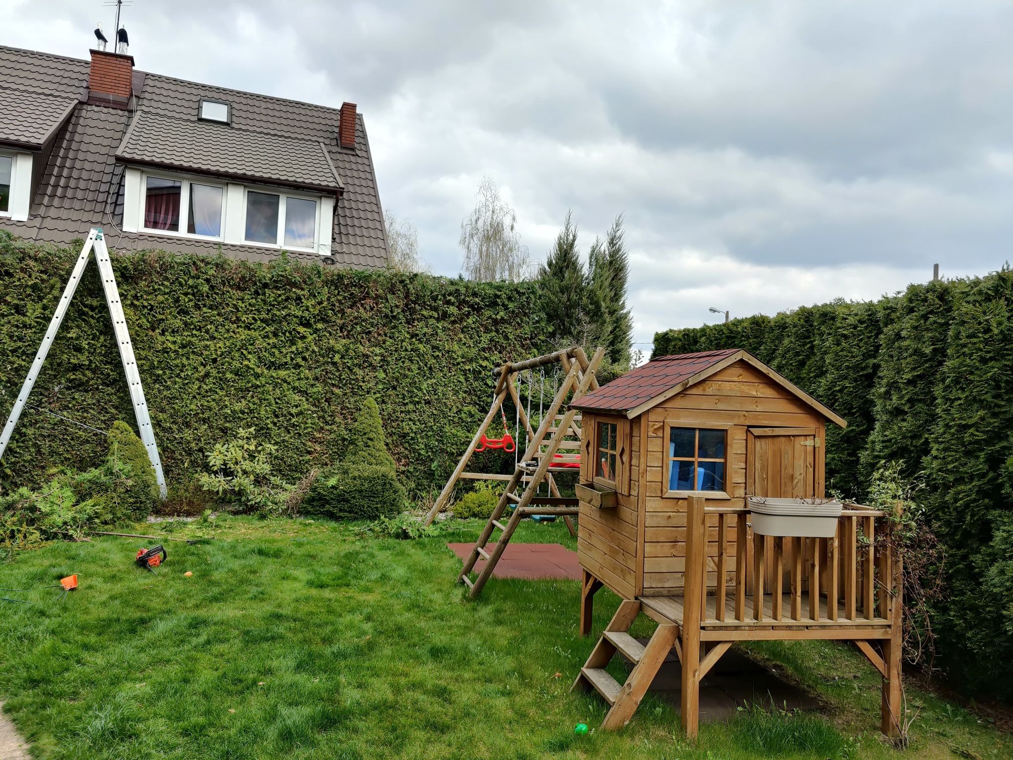 Przydomowy ogród z drewnianym domkiem dla dzieci na podwyższeniu, huśtawką, żywopłotem z tuj i drabiną opartą o ścianę domu, po przycięciu gałęzi.