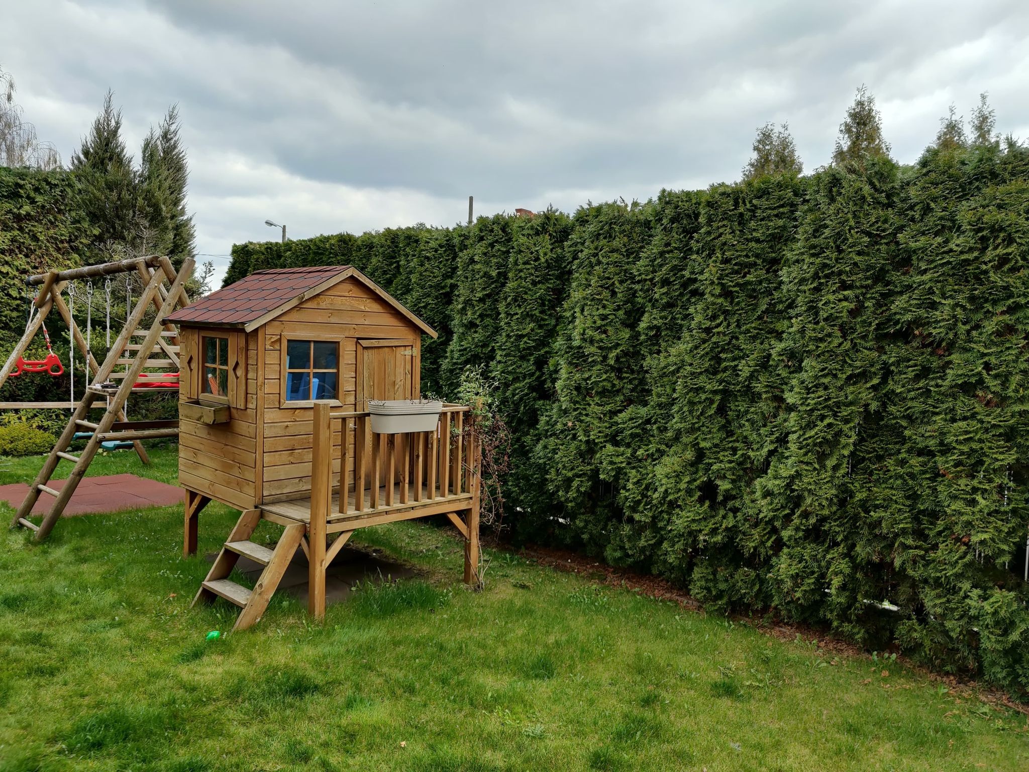 Drewniany domek dla dzieci z czerwonym dachem, umieszczony na podwyższeniu z schodkami, obok huśtawki i wysokiego, gęstego żywopłotu z tui na trawniku w pochmurny dzień.