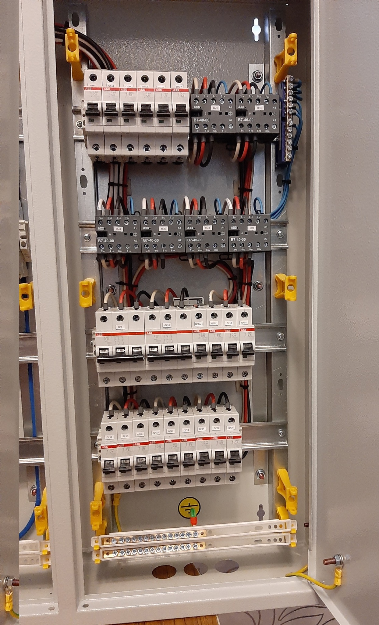 Otwarta skrzynka elektryczna z bezpiecznikami, przekaźnikami i przewodami, widoczne szyny montażowe i żółte elementy mocujące, uziemienie z zielonym akcentem.