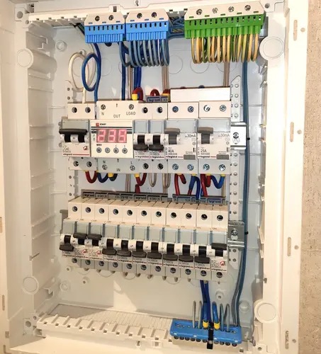 Otwarta skrzynka elektryczna z zainstalowanymi bezpiecznikami, przewodami w kolorach niebieskim, zielonym, czerwonym i żółtym, oraz licznikiem elektronicznym z czerwonym wyświetlaczem.