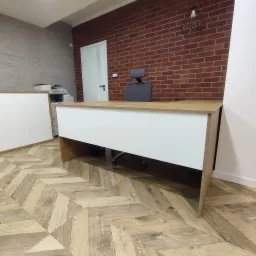 Nowoczesne biuro z drewnianą podłogą w jodełkę, biurkiem z białym frontem i blatem w kolorze drewna, ceglaną ścianą z dekoracyjną lampą i szarą ścianą w tle.