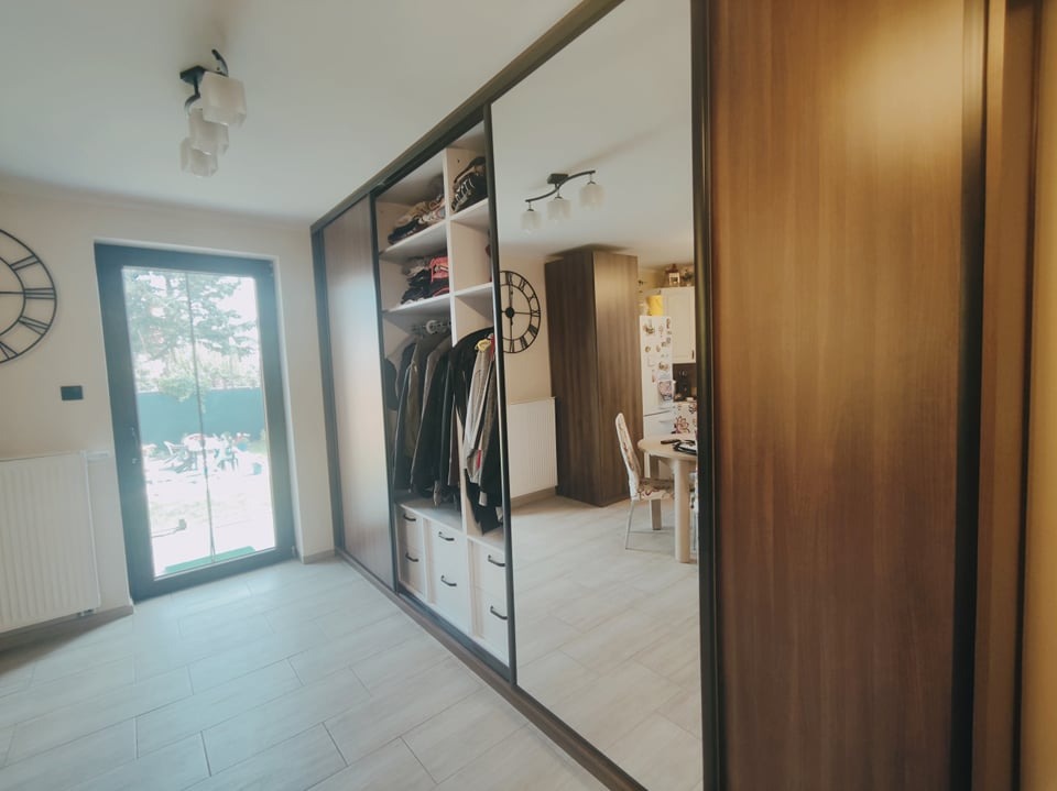 Szafa wnękowa z przesuwnymi drzwiami lustrzanymi, widoczne ubrania na wieszakach i półkach, jasne wnętrze, w odbiciu lustrzanym widok na kuchnię z zegarem na ścianie.