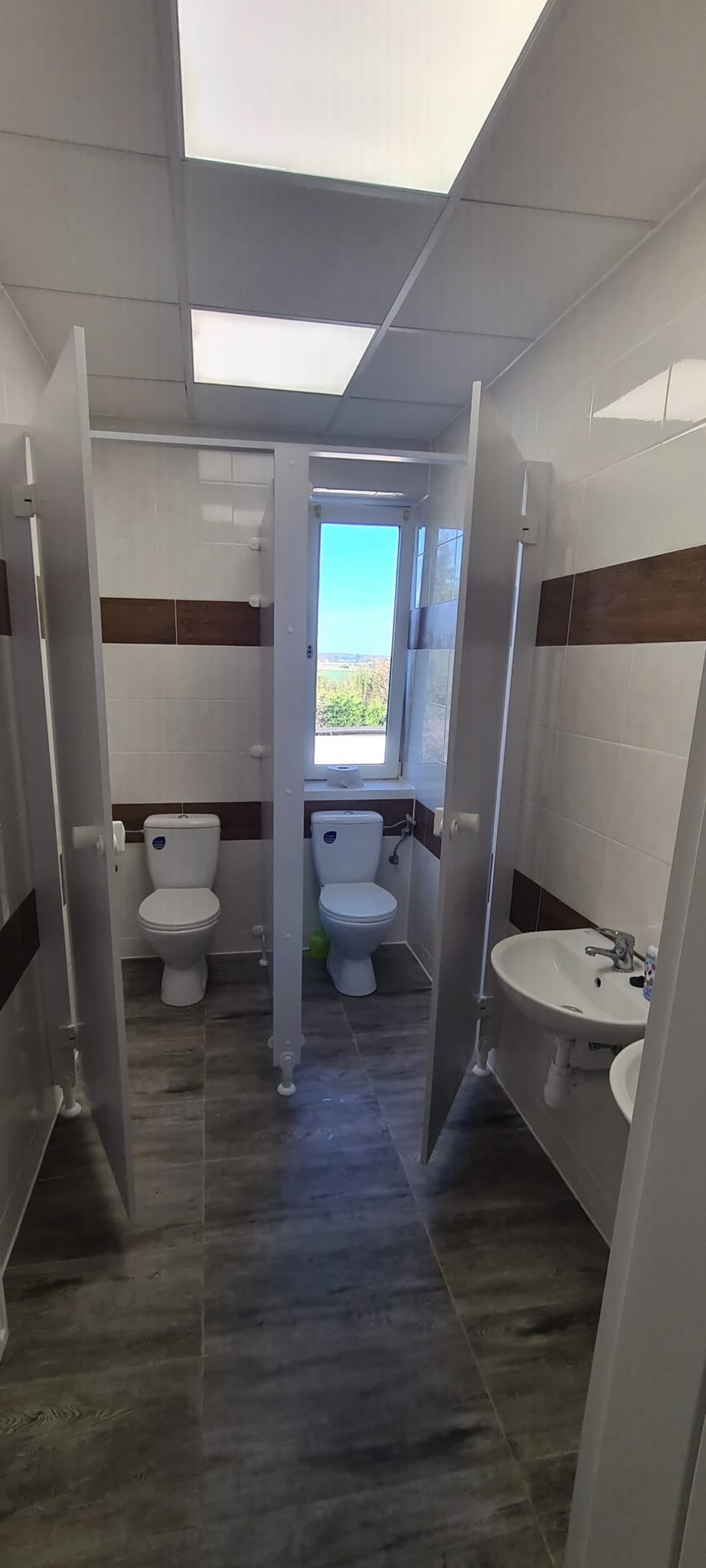 Wnętrze publicznej toalety z kabinami, widoczne dwie toalety, umywalki, szare płytki podłogowe imitujące drewno, biało-beżowe płytki ścienne z brązowym pasem, okno z widokiem na zieleń.